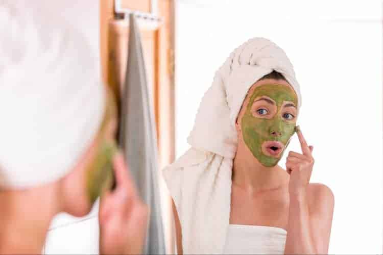 woman staring at her reflection in mirror with matcha green tea mask on her face